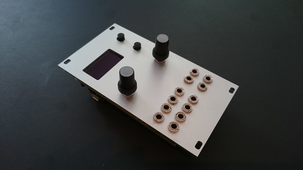 Ornament and Crime Eurorack module seen at an angle, with OLED display, two encoders and a grid of jacks on a silver panel
