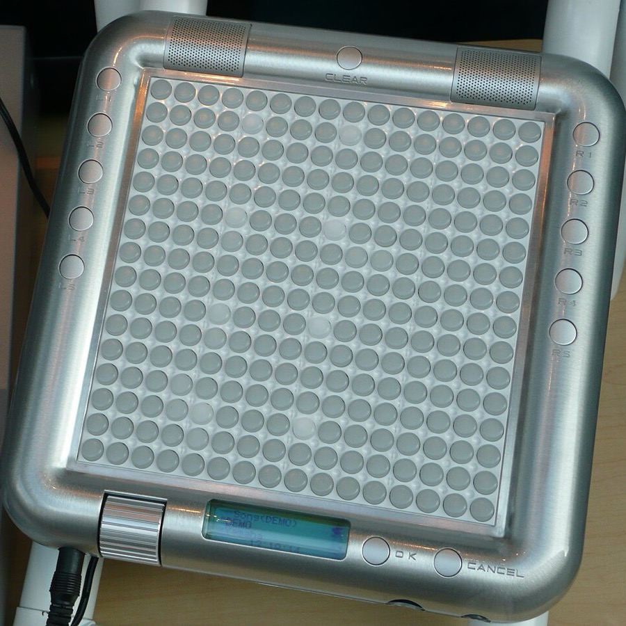 Silver Yamaha Tenori-on with its 16 by 16 button grid, side buttons and small display, cropped from a photo beside a mixing desk